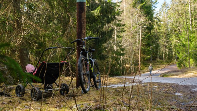 Sykkel og barnevogn parkert i Oslomarka. Dame på tur med hund i bakgrunnen.