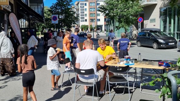 Flere mennesker som oppholder seg på et fortau. Det er fint vær. To mennesker spiller sjakk rundt et bord med flere brettspill.