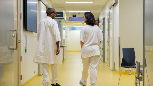 To helsearbeidere i arbeidstøy går nedover en korridor.