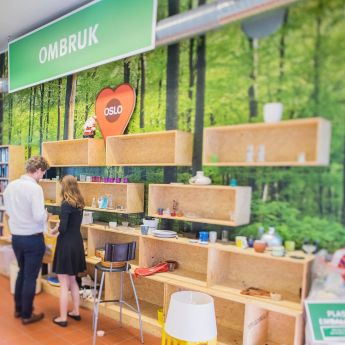 En kvinne og en mann står foran hyllene i en minigjenbruksstasjon og kikker på gjenstandene i hyllene.