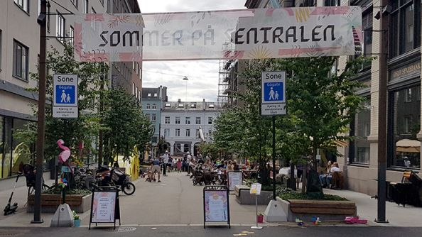 Gåate med trær og benker på begge sider. Banner med påskriften "sommer på sentralen" mellom to stolper.
