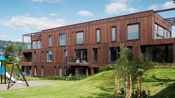Bygg på tre etasjer i tre. Høye vinduer og terrasse med glassgjerde og overbygg.