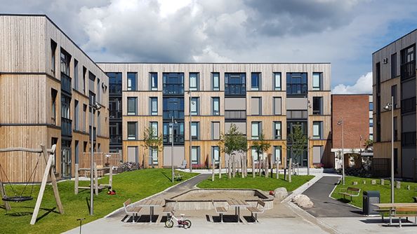 Tre fireetasjes blokker med smalt trepanel. Lekeplass og benker mellom blokkene.