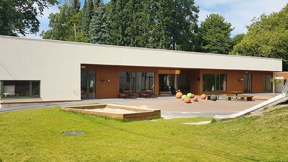 Lavt, flatt bygg med hvit utkraving over førsteetasje med trepanel og store vinduer. Lekeplass i forgrunnen.