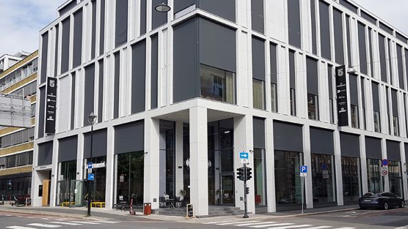 Treetasjes kontorbygg med store vindusflater, og struktur i fasaden. Andre etasje stikker ut over næringslokalene i første etasje.