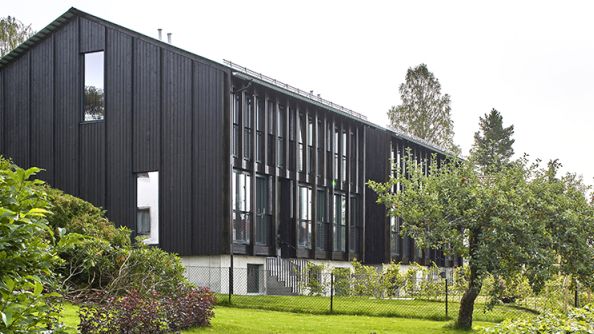 Hus i grønn hage. Smalt panel og skråtak. To høye vinduer på kortsiden, tette vinduer på langsiden.