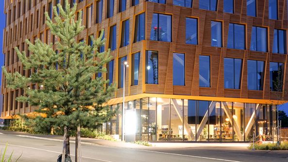 Kveldsbilde av de nederste etasjene av et stort kontorbygg i tre. Glassfasade i første etasje, og en utkraging fra andre etasje og oppover.