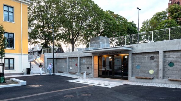 Hjørne av en skolegård, med et eldre gult bygg på venstre side, og et nyere lavere steinbygg på høyre side.