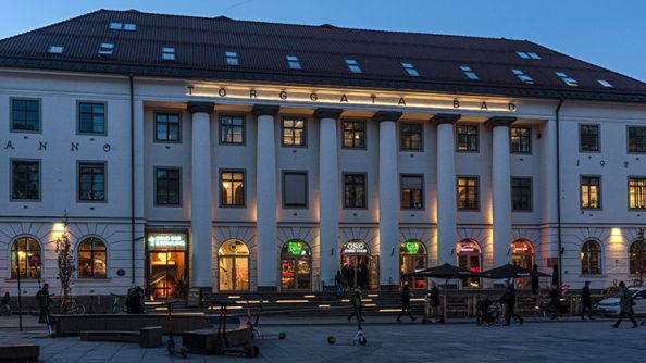 Stort sentrumsbygg, med søyler i front, og skråtak med takvinduer. Opplyst "Torggata bad" over søylene.