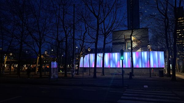 Byggegjerdet til Oslo Spektrum om kvelden, med blå‑rosa lys og trær i silhuett foran.