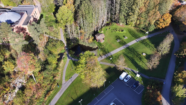 Flyfoto av park ved vann med sti, trær og liten dam nær en bygning til venstre.