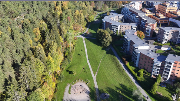Flyfoto av grøntområde mellom skog og boligblokker. Stier går gjennom gresset, med bålplass og åpne oppholdsarealer.