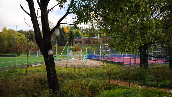Lekeområde med klatrestativ i sand ved siden av fotballbane og basketbane. Trær og grønt landskap rammer inn området.