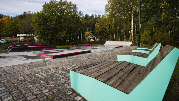 Skatepark med ramper og rails ved siden av sittebenker i et grønt område. Trær og sti omgir plassen, og underlaget er vått etter regn.