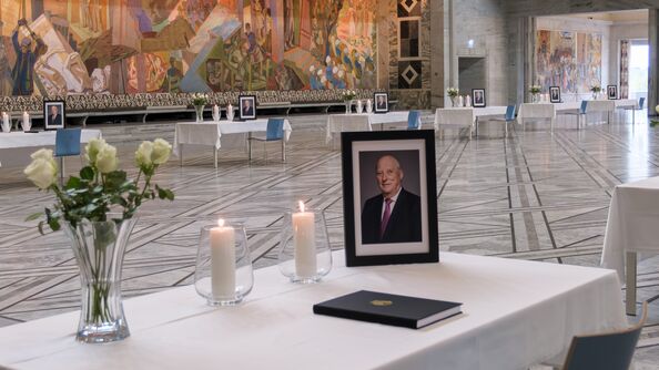 Et bord med hvit duk, lys, blomster og portrett av kong Harald V.På bordet ligger en kondolanseprotokoll klar til å åpnes og signeres.