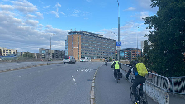 Syklister sykler på et fortau langs en bro med biltrafikk. Det finnes ikke et tydelig avgrenset sykkelfelt, og syklister og gående deler arealet.