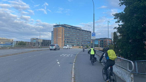 Syklister sykler på et fortau langs en bro med biltrafikk. Det finnes ikke et tydelig avgrenset sykkelfelt, og syklister og gående deler arealet.