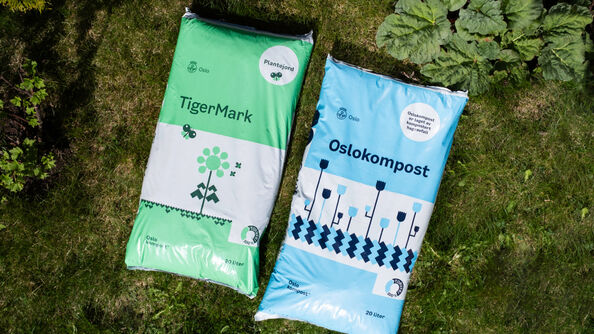 To sekker med jordprodukter, TigerMark og Oslokompost, ligger på gress ved siden av grønne planter. Produktene er laget av kompostert hage- og matavfall og brukes til planting og hagearbeid.