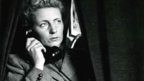 Gammelt svart-hvitt fotografi av Else Wendel som snakker i telefonen mens hun står mellom gardiner i et avskjermet område.