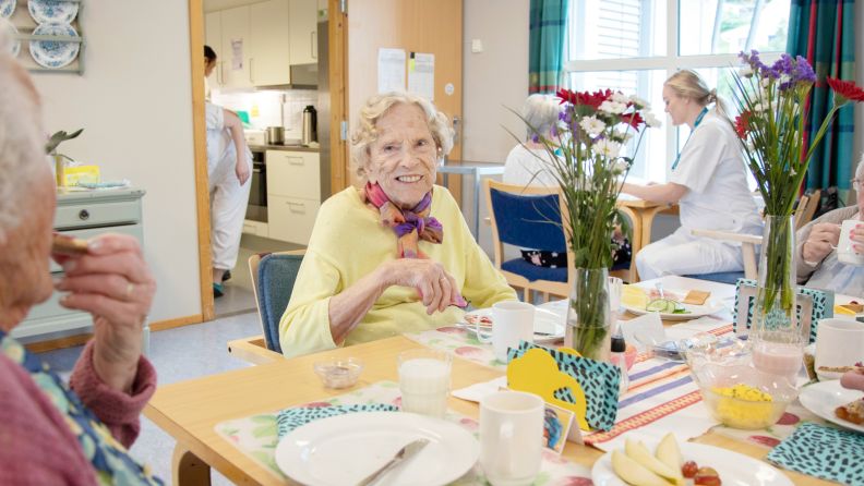 Beboere smiler og koser seg sammen med felles frokost med blomsterpynt på bordet.