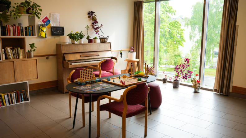 Et rom med store vinduer, et piano og et bord i midten av rommet med flere brettspill. 