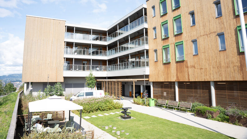 Et tre-etasjers helsehusbygg i lyst treverk. Hver etasje har grønne persienner og balkong.