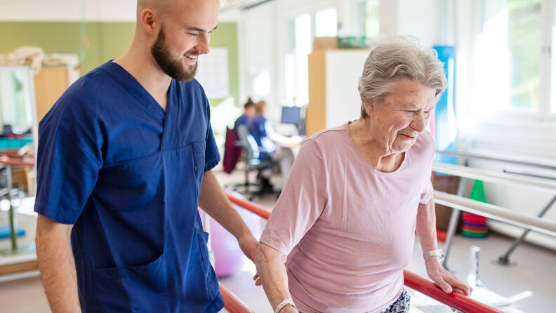 Fysioterapaut hjelper smilende pasient med gåtrening i helsehusets rehabiliteringsrom.