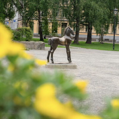 Skulptur av en hest på en sokkel i en park, med trær og en bygning i bakgrunnen.