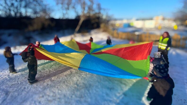 Flere barn og en voksen leker med en fargerik fallskjerm ute i snøen