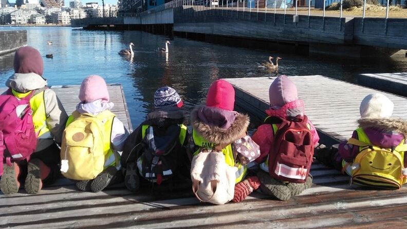 Seks barn med sekk og parkdress på tur til Sørenga. Svaner og Bjørvika i bakgrunn. 