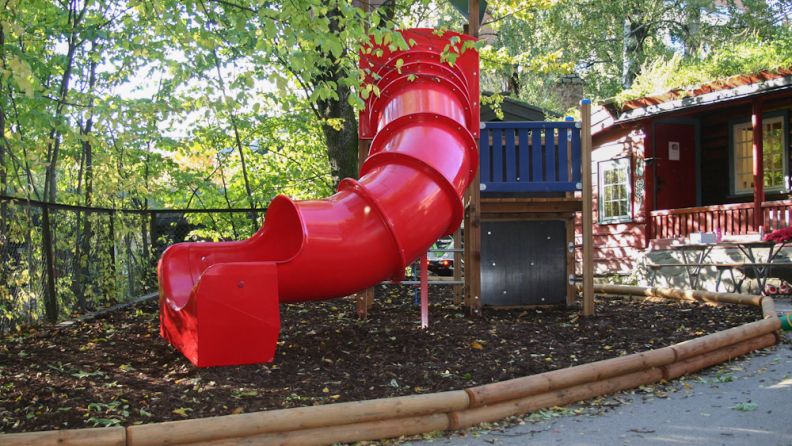 Uteområde med sklie, klatrestativ som står på jord, med gjerde og trær rundt