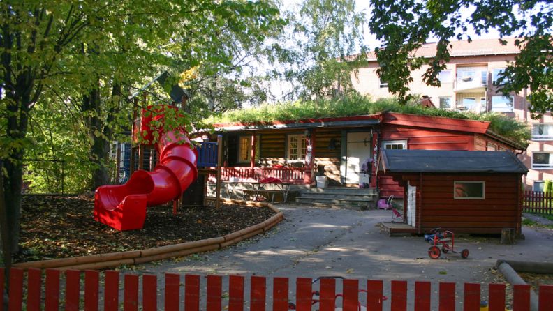 Uteområde med sklie, lekehus og trær rundt, rødmalt barnehage i bakgrunnen