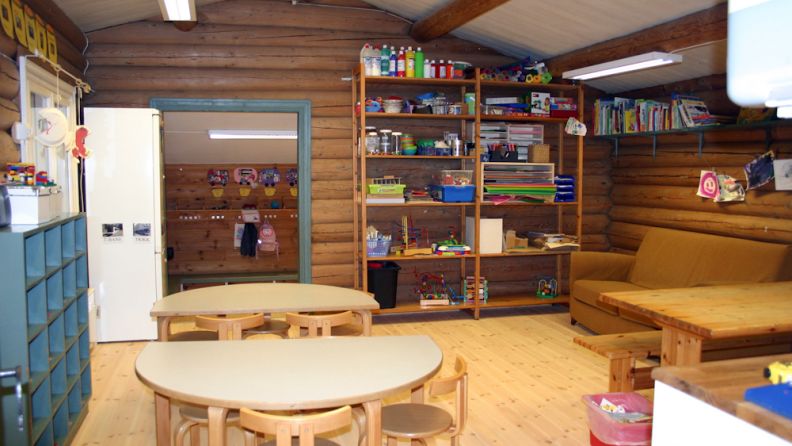 Tømmerkledd rom, med 3 bord stoler sofa, hyller med leker og bøker