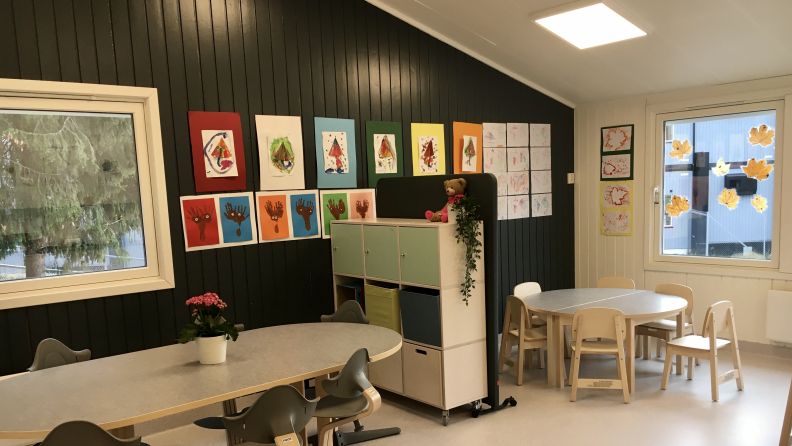 Inne i en avdeling med bord, leker og tegninger på veggen. 