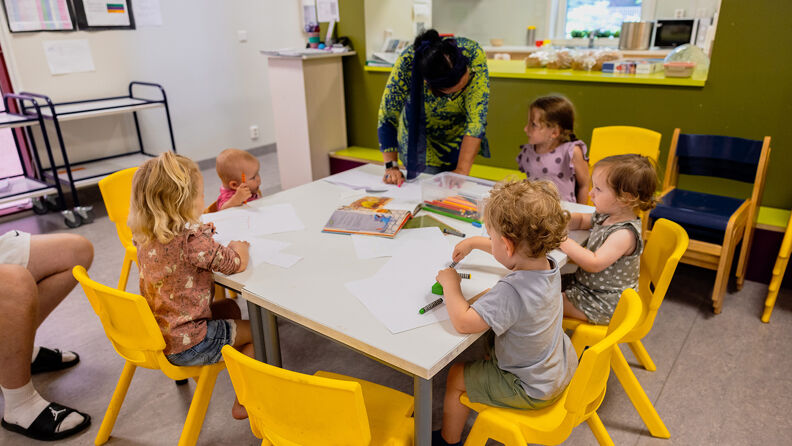 Små barn sitter rundt et bord og tegner. En voksen hjelper til.